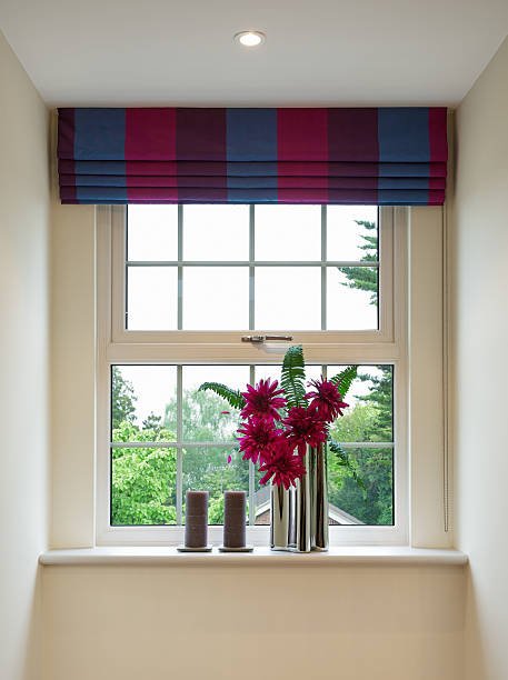 a Geogian style sash window with colourful blinds overhead and complementary red flowers in a modern chrome vase below, next to two brown candles. In the background is a bright view to a garden with lots of trees.