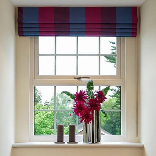 a Geogian style sash window with colourful blinds overhead and complementary red flowers in a modern chrome vase below, next to two brown candles. In the background is a bright view to a garden with lots of trees.